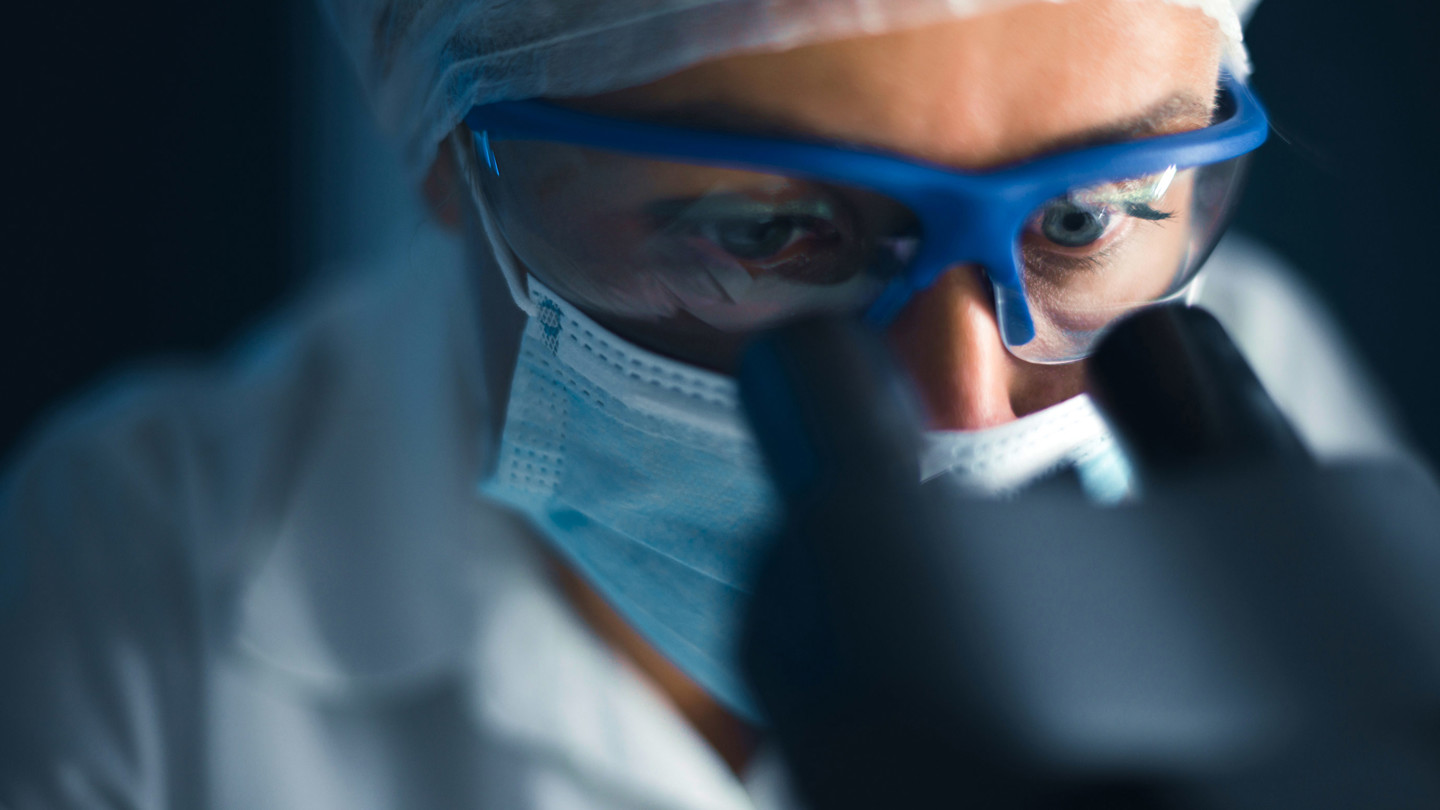 Person in mask and safety goggles looking through a microscope